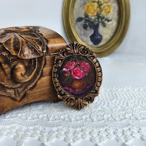 May include: A small, oval-shaped painting of pink roses in a vase, set in a decorative bronze frame. The frame is ornate with a detailed, vintage design. A wooden box with carved floral details is to the left, and a framed painting of yellow roses is in the background.