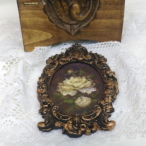 May include: A small, oval-shaped painting of white roses in a gold-colored frame. The painting is set against a dark purple background.