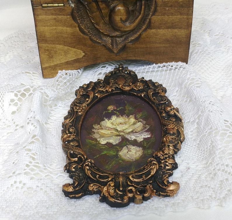 May include: A small, oval-shaped painting of white roses in a gold-colored frame. The painting is set against a dark purple background.