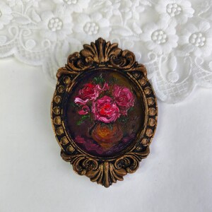 May include: An oval-shaped, bronze-colored frame with an oil painting of pink roses in a vase. The painting has a dark background with visible brushstrokes. The frame has ornate detailing and a vintage aesthetic.