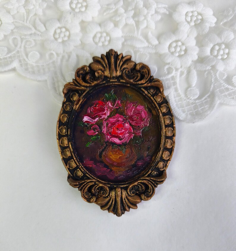 May include: An oval-shaped, bronze-colored frame with an oil painting of pink roses in a vase. The painting has a dark background with visible brushstrokes. The frame has ornate detailing and a vintage aesthetic.