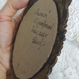 May include: A close-up of the back of a small oval-shaped painting in a gold ornate frame. The painting is on cardboard and is signed "Peonies Oil/cardboard Year: 2024  Rhuefi."