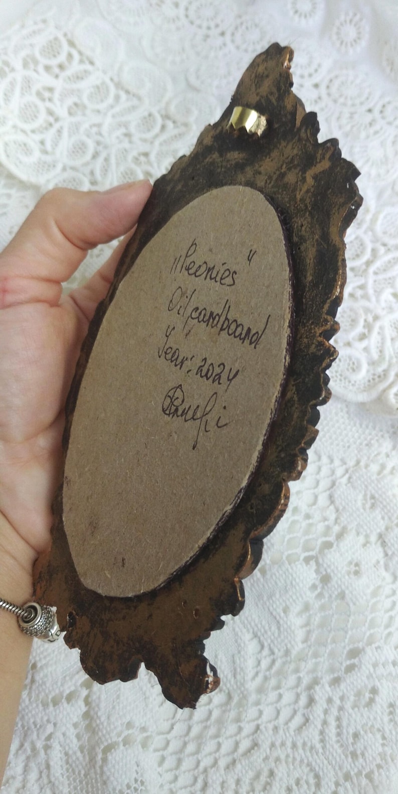 May include: A close-up of the back of a small oval-shaped painting in a gold ornate frame. The painting is on cardboard and is signed "Peonies Oil/cardboard Year: 2024  Rhuefi."