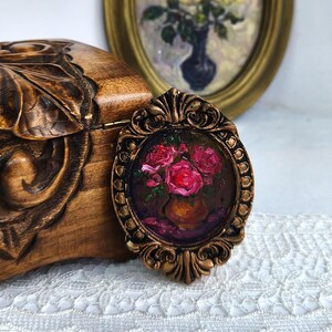 May include: A small, oval-shaped painting in a bronze-colored ornate frame. The painting depicts a vase of red roses. The frame is set against a wooden box with carved floral details and a vintage painting in the background.