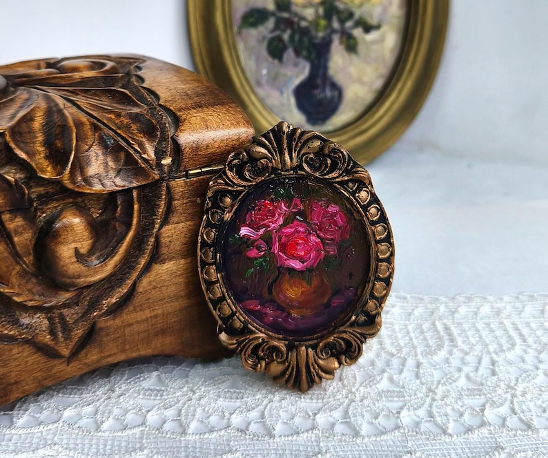 May include: A small, oval-shaped painting in a bronze-colored ornate frame. The painting depicts a vase of red roses. The frame is set against a wooden box with carved floral details and a vintage painting in the background.