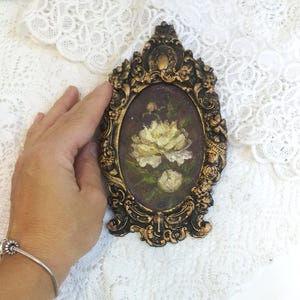 May include: A small oval painting of a white rose in a gold ornate frame. The painting is set against a dark purple background.