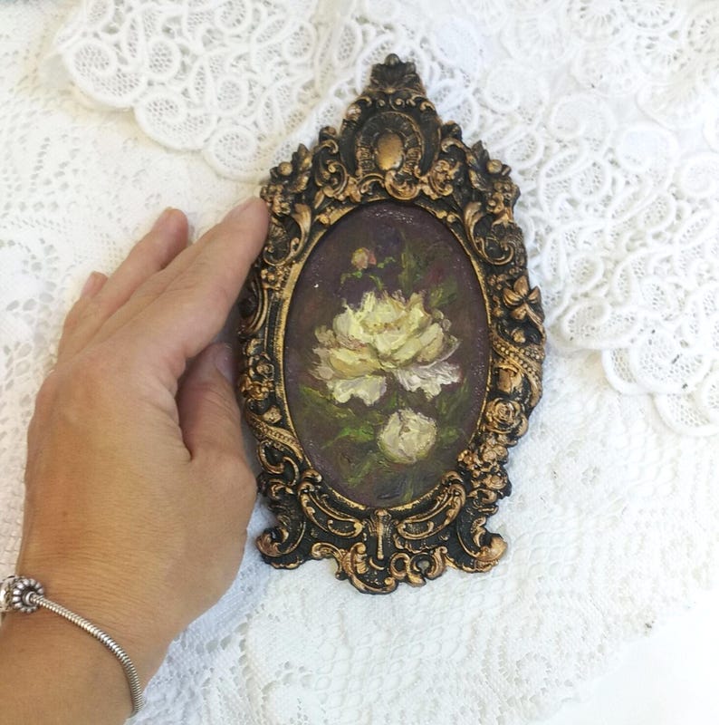 May include: A small oval painting of a white rose in a gold ornate frame. The painting is set against a dark purple background.