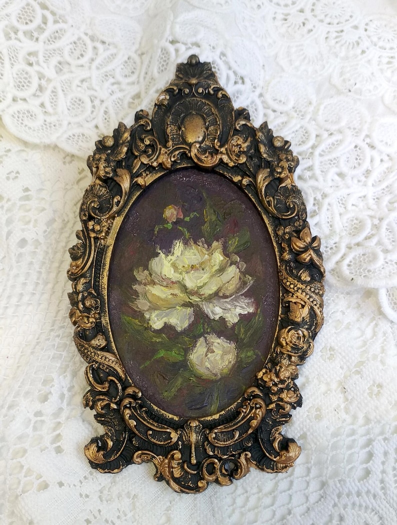 May include: A white flower painting in an ornate gold and black oval frame. The painting depicts a white flower with green leaves on a dark purple background.