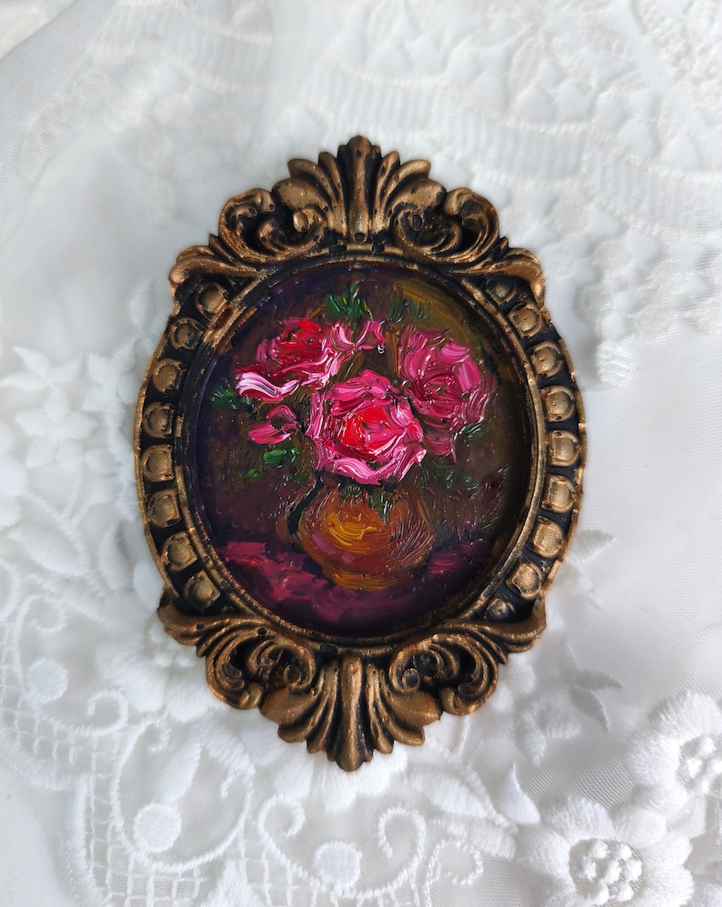 May include: Oval-shaped oil painting of pink roses in a vase, set in a decorative gold-colored frame. The artwork features a dark background and a textured, impressionistic style, evoking a classic aesthetic.