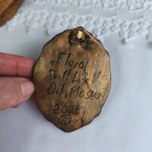 May include: A bronze-colored, oval-shaped plaque with the inscription "Floral Still Life" and the artist's name, "O.J. Plaster", along with the year 2023. The plaque has a small metal loop for hanging.