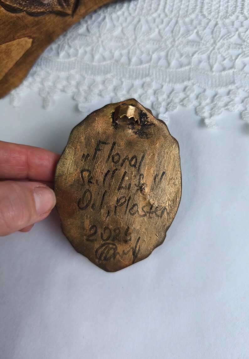 May include: A bronze-colored, oval-shaped plaque with the inscription "Floral Still Life" and the artist's name, "O.J. Plaster", along with the year 2023. The plaque has a small metal loop for hanging.