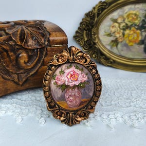 May include: A small, oval-shaped painting in a decorative gold frame. The painting depicts a vase of pink roses. A carved wooden box and another framed painting are in the background. The painting is on a white lace surface.