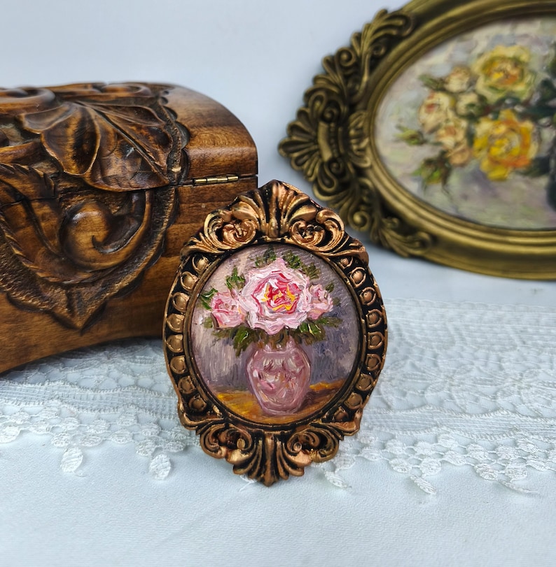May include: A small, oval-shaped painting in a decorative gold frame. The painting depicts a vase of pink roses. A carved wooden box and another framed painting are in the background. The painting is on a white lace surface.