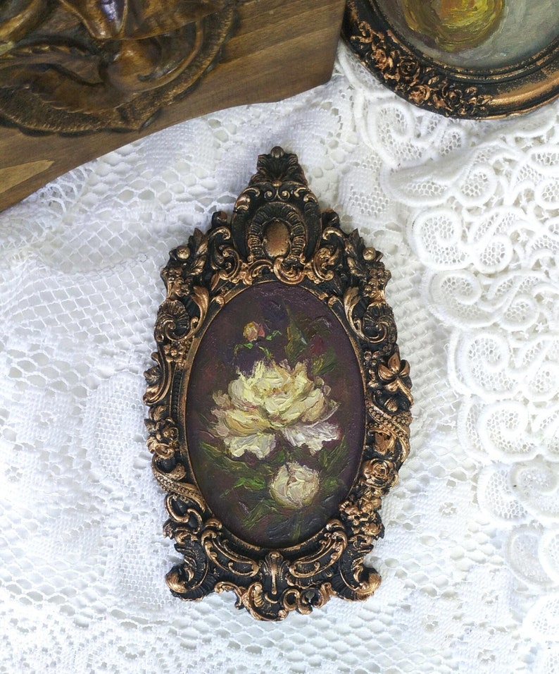 May include: A small oval painting of white peonies in a dark brown ornate frame. The frame is gold-gilded and has a detailed design.