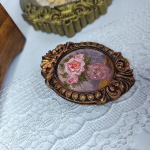 May include: An oval-shaped, antique-style brooch with a miniature painting of pink roses and a pink object. The brooch has a detailed, ornate gold-colored frame. The background is a light blue and purple color. The brooch is placed on a white lace surface.