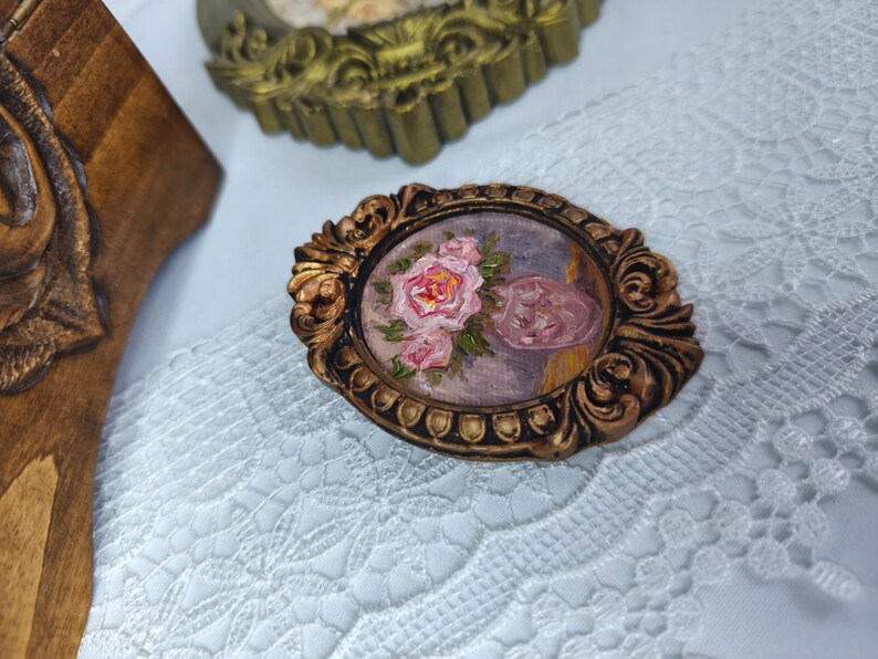 May include: An oval-shaped, antique-style brooch with a miniature painting of pink roses and a pink object. The brooch has a detailed, ornate gold-colored frame. The background is a light blue and purple color. The brooch is placed on a white lace surface.