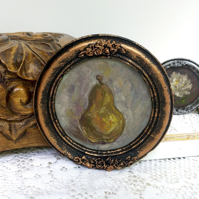 Puede incluir: Una peque&ntilde;a pintura redonda de una pera en un marco dorado y negro. La pera est&aacute; pintada en un estilo realista con tonos de verde, amarillo y marr&oacute;n. El fondo es un morado suave y apagado.