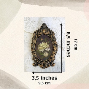 May include: A gold-toned oval frame with ornate details, featuring a painting of white flowers on a dark background. The frame measures 6.5 inches tall and 3.5 inches wide.