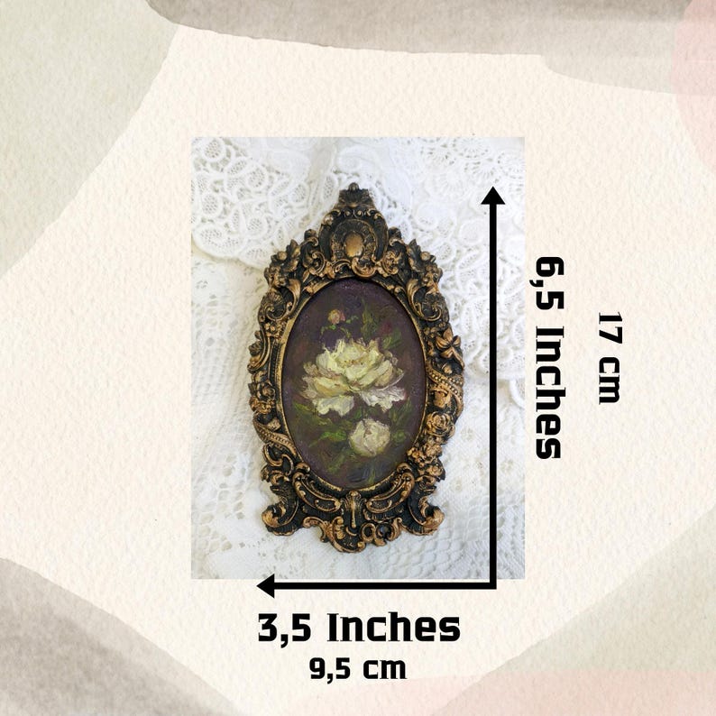 May include: A gold-toned oval frame with ornate details, featuring a painting of white flowers on a dark background. The frame measures 6.5 inches tall and 3.5 inches wide.