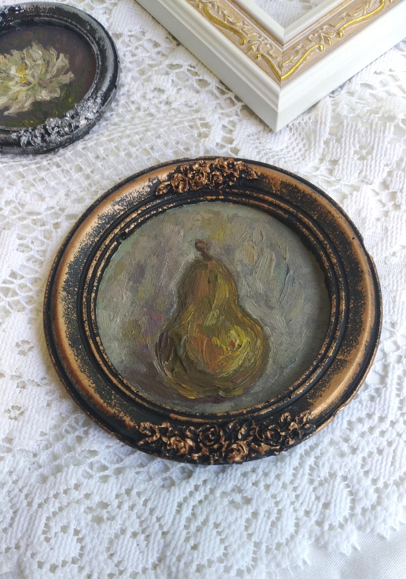 Puede incluir: Una pintura al &oacute;leo enmarcada de una pera en tonos verdes y amarillos, sobre un fondo tenue. El marco es negro con detalles dorados y un dise&ntilde;o floral decorativo.
