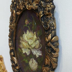May include: A small oval painting of white flowers in a gold and black ornate frame.