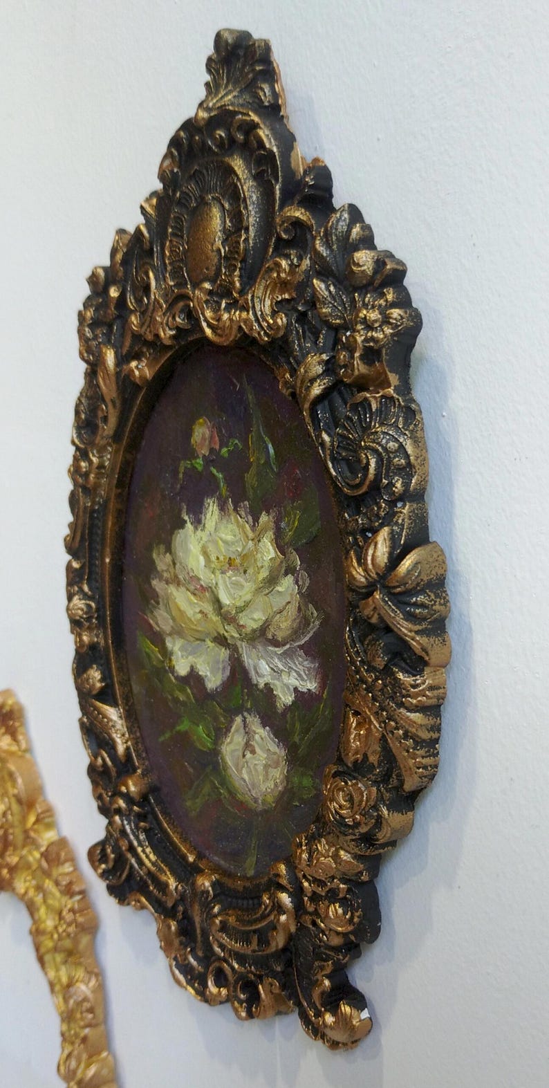 May include: A small oval painting of white flowers in a gold and black ornate frame.