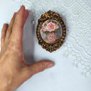 May include: An oval-shaped, antique-style, gold-colored frame with a miniature painting of pink roses in a vase. The painting is set against a muted background. The frame has ornate detailing.