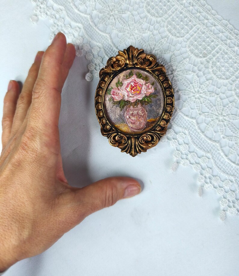 May include: An oval-shaped, antique-style, gold-colored frame with a miniature painting of pink roses in a vase. The painting is set against a muted background. The frame has ornate detailing.