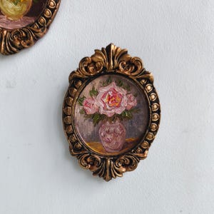 May include: A small, oval-shaped, framed painting featuring a bouquet of pink roses in a vase. The painting is set within a decorative, ornate, gold-colored frame. The artwork has a vintage aesthetic.
