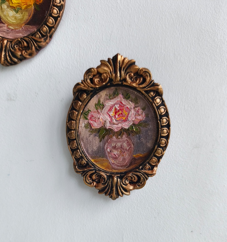 May include: A small, oval-shaped, framed painting featuring a bouquet of pink roses in a vase. The painting is set within a decorative, ornate, gold-colored frame. The artwork has a vintage aesthetic.