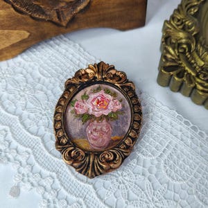May include: An oval-shaped, ornate, bronze-colored frame surrounds a small painting of pink roses in a pink vase. The painting is set against a backdrop of purple and yellow hues. The frame has a detailed, textured design.