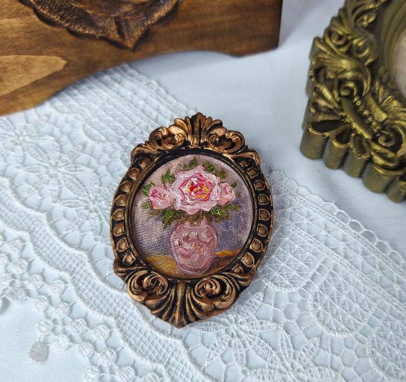 May include: An oval-shaped, ornate, bronze-colored frame surrounds a small painting of pink roses in a pink vase. The painting is set against a backdrop of purple and yellow hues. The frame has a detailed, textured design.