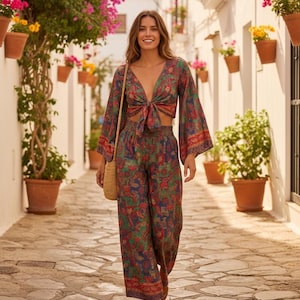 May include: A woman in a two-piece floral print outfit, featuring a tie-front top and wide-leg trousers. The ensemble has a red, green, and blue paisley pattern. She carries a woven shoulder bag and walks down a cobbled street.