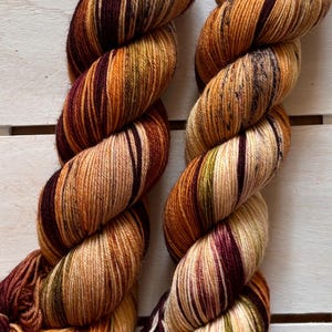Op de afbeelding: Twee strengen handgeverfd garen in een rijk kleurenpalet van bordeauxrood, goud en olijfgroen. Het garen is gedraaid in een touwachtige vorm, die de kleurvariaties laat zien. De strengen liggen op een wit houten oppervlak.