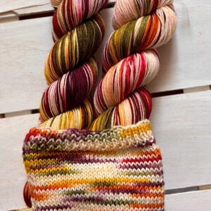 Könnte beinhalten: Zwei Stränge bunter Wolle mit gestrickter Manschette. Das Garn ist eine Mischung aus Rot, Orange, Gelb, Grün und Lila. Die Manschette ist eine Mischung aus den gleichen Farben. Ein Etikett ist am Garn befestigt.