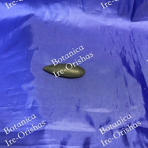 May include: A small, dark gray, smooth, oval-shaped stone with the text "Botanica Ire-Orishas" printed on it. The stone is sitting on a blue fabric surface.