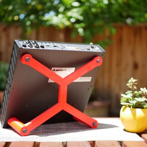 May include: A black electronic device with a red stand. The device has multiple ports and buttons on the back. The stand is made of plastic and has an X-shaped design.