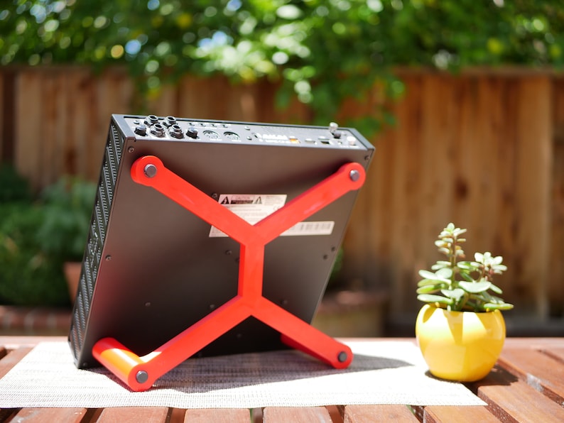 May include: A black electronic device with a red stand. The device has multiple ports and buttons on the back. The stand is made of plastic and has an X-shaped design.