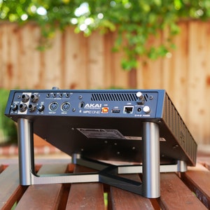May include: Black Akai MPC One music production studio with silver metal stand legs. The rear of the device shows various ports and connections, including USB, MIDI, and power.