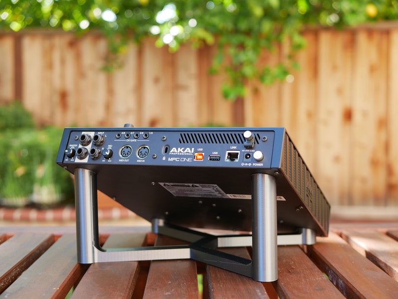 May include: Black Akai MPC One music production studio with silver metal stand legs. The rear of the device shows various ports and connections, including USB, MIDI, and power.