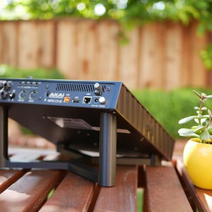 May include: Black Akai MPC One music production studio with a stand and a yellow pot with a green plant on a wooden table.