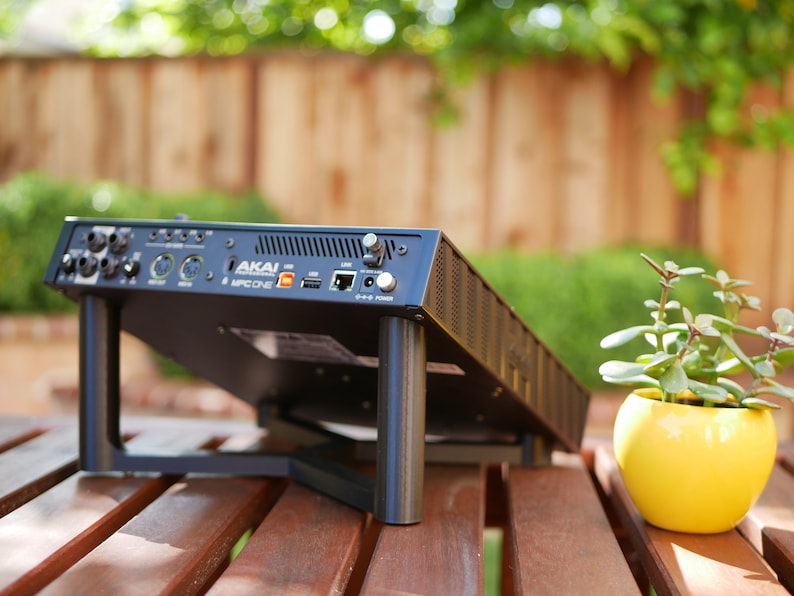 May include: Black Akai MPC One music production studio with a stand and a yellow pot with a green plant on a wooden table.