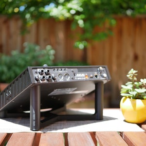 May include: Black audio interface with multiple ports and connections, sitting on a wooden table with a small yellow planter with a green succulent plant.