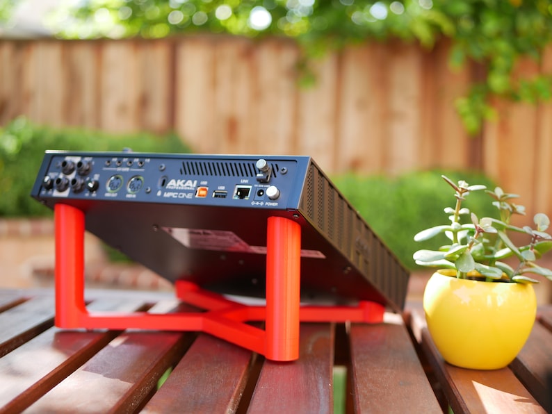 May include: A black Akai MPC One music production studio with a red stand, sitting on a wooden table. A small potted plant with green leaves is on the table to the right of the stand.