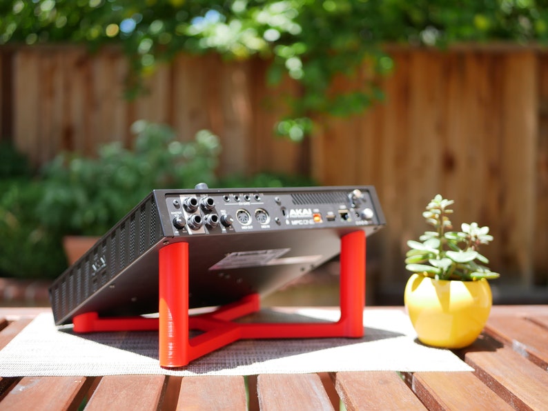 May include: A black music production interface with a red stand, the interface has a label that says "AKAI LPK25". The interface is sitting on a wooden table with a small yellow pot with a green plant.