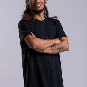 Peut inclure: Un homme aux cheveux longs et foncés porte un t-shirt noir et un short blanc. Il sourit et a les bras croisés.