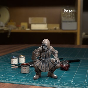 Peut inclure: Une figurine miniature d'un personnage portant une tenue blindée à capuche, accroupi avec une main levée dans un geste. La figurine est peinte en bronze et présente un design détaillé. La figurine est posée sur un tapis de coupe vert avec un dé à 20 faces.