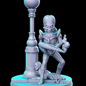 Op de afbeelding: Een grijs, gedetailleerd beeldje van een alien met een stralenpistool, staand boven een neerliggende menselijke figuur. De alien draagt een futuristisch pak en heeft een groot hoofd met een schedel-achtig gezicht. De scène speelt zich af op een grijze, ronde basis met een lantaarnpaal op de achtergrond.