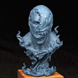 Puede incluir: Un busto de un personaje impreso en 3D en azul con un rostro humano y un simbionte que cubre la mitad de su rostro. El simbionte tiene dientes afilados y una superficie texturizada. El busto está sobre una base de madera.