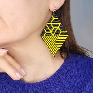 Puede incluir: Un par de pendientes geométricos negros y amarillos. Los pendientes están hechos de pequeñas cuentas y tienen forma de diamantes. Los pendientes cuelgan de los lóbulos de las orejas de una persona que lleva un suéter azul.